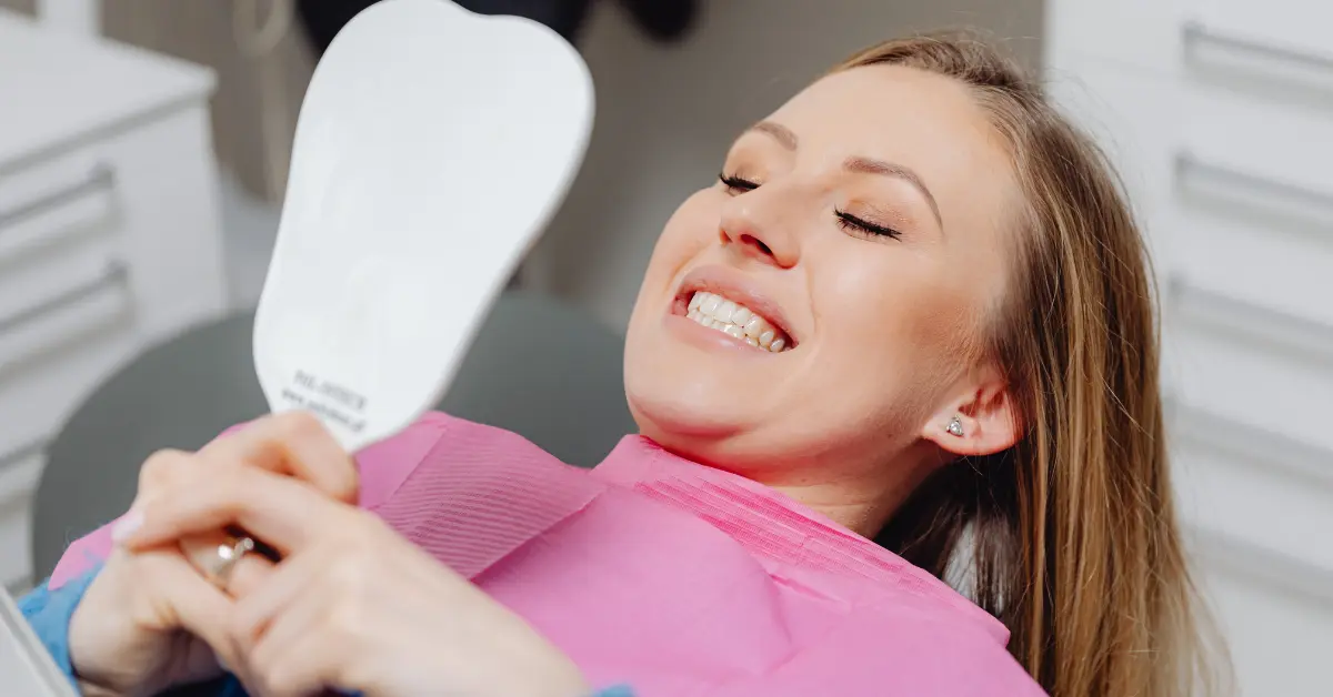 A smiling woman in a dental chair wearing a pink bib admiring her teeth in a hand mirror after a professional dental cleaning