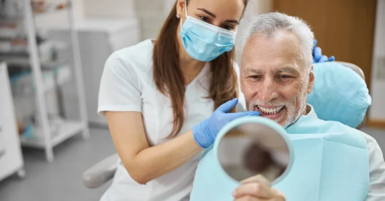 Smiling senior man examining his teeth in a handheld mirror while a dental professional checks his smile during a dental appointment.